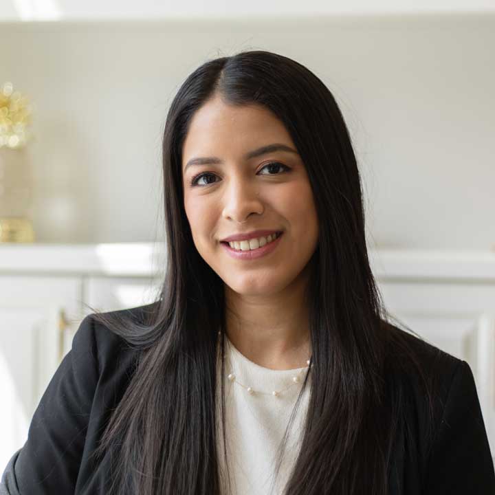 Formal headshot of Adzuira Musule Palacios, smiling playfully at the camera, with long ironed hair, wearing a formal black jacket and a cute pearl neacklace over a cream blouse. She is sitting on an off-frame couch in a brightly lit room. The background contains a bookshelf with one yellow item split in half by the edge of the frame, but detail is missing as it is faded with a bokeh effect.